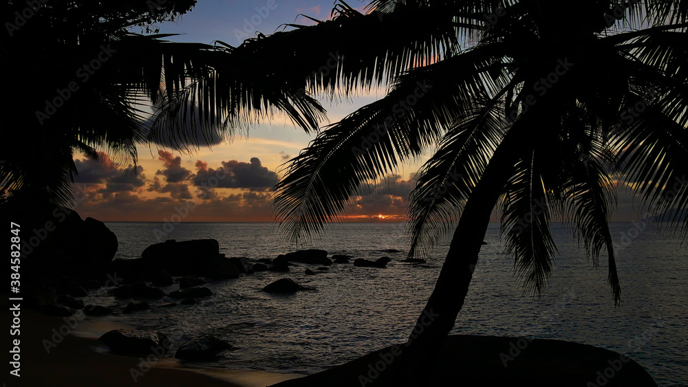 Beautiful sunset with sun disappearing in the clouds over the horizon of the ocean on Tusculum Beach, Mahe island, Seychelles with the silhouettes of palm trees and granite rocks.