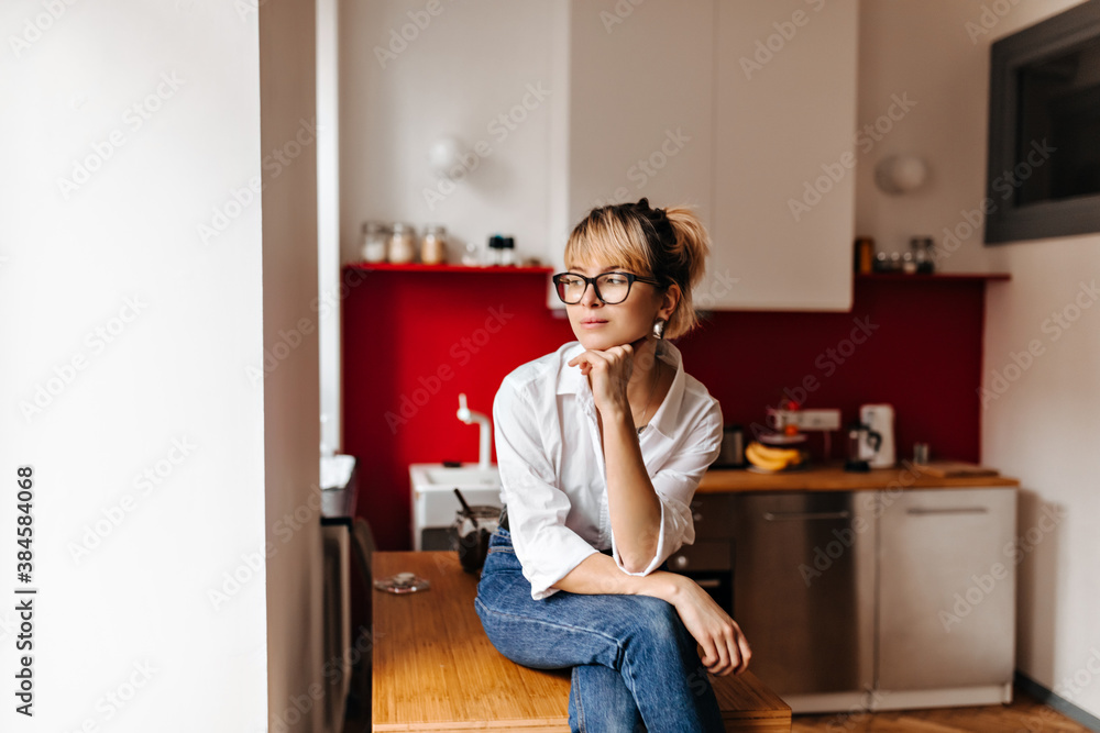 Pensive girl with tattoo sitting on wooden table. Indoor shot of dreamy woman wearing white shirt and jeans. Pensive girl with tattoo sitting on wooden table. Indoor shot of dreamy woman wearing white shirt and jeans.