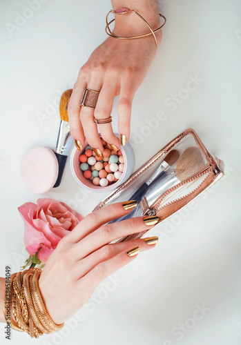 woman hands with golden manicure and many rings holding brushes, makeup artist stuff stylish, pure closeup pink flower rose among cosmetic for makeup