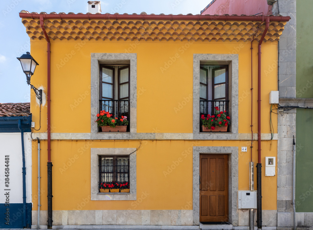 Traditional Spanish house. Old building restored, facade painted in yellow. Windows and balcony framed with grey stone. Wooden door. Red geraniums decorating the windows. Asturias, North of Spain