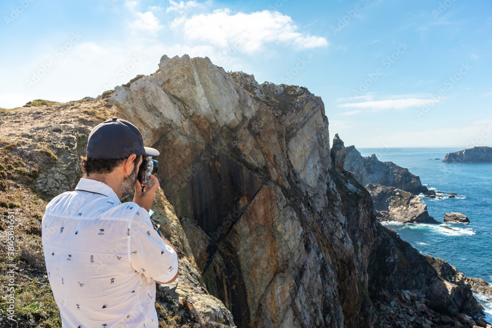 Fototapeta premium Male tourist taking pictures of the ocean on top of a high cliff. He is using a retro style photography camera. Summer time. Cape Penas (Cabo de Peñas) is the most northerly point of Spain, Asturias