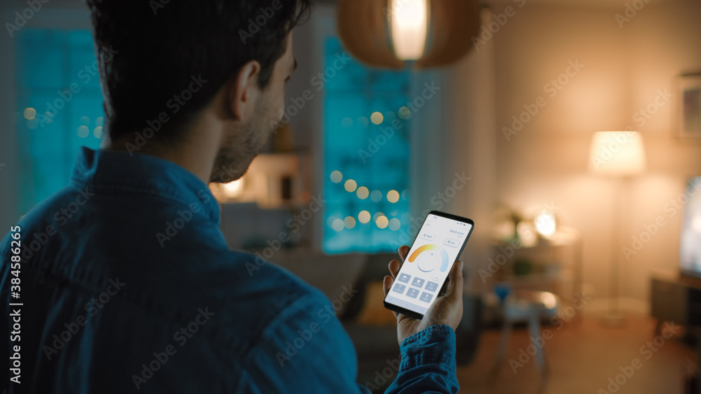 Young Handsome Man is Setting up Comfortable Bedroom Conditions with Voice Command to Smart Home Application on His Smartphone. It's a Cozy Evening in Technologically Advanced Living Room. Young Handsome Man is Setting up Comfortable Bedroom Conditions with Voice Command to Smart Home Application on His Smartphone. It's a Cozy Evening in Technologically Advanced Living Room.