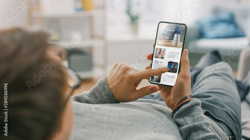 Obraz na plátně Young Man at Home is Lying on a Sofa and Using Smartphone for Scrolling and Reading News about Technological Breakthroughs