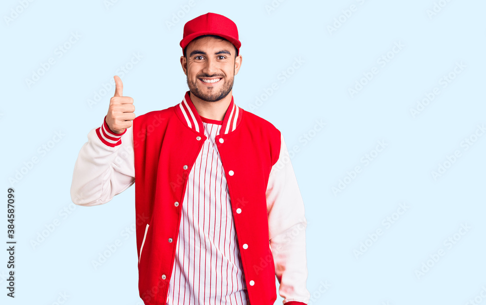 Young handsome man wearing baseball jacket and cap smiling happy and positive, thumb up doing excellent and approval sign Young handsome man wearing baseball jacket and cap smiling happy and positive, thumb up doing excellent and approval sign