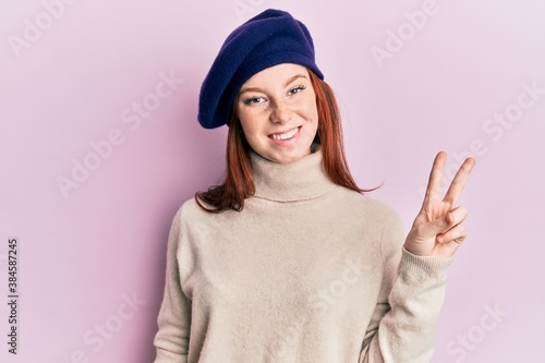 Young red head girl wearing french look with beret smiling with happy face winking at the camera doing victory sign. number two. © Krakenimages.com