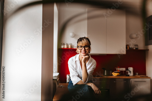 Selective focus of happy young woman in glasses. Blissful blonde girl sitting in kitchen and looking at camera.