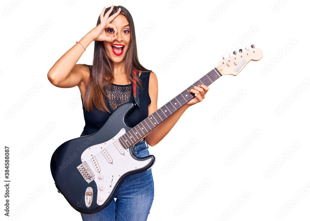 Young hispanic woman playing electric guitar smiling happy doing ok sign with hand on eye looking through fingers