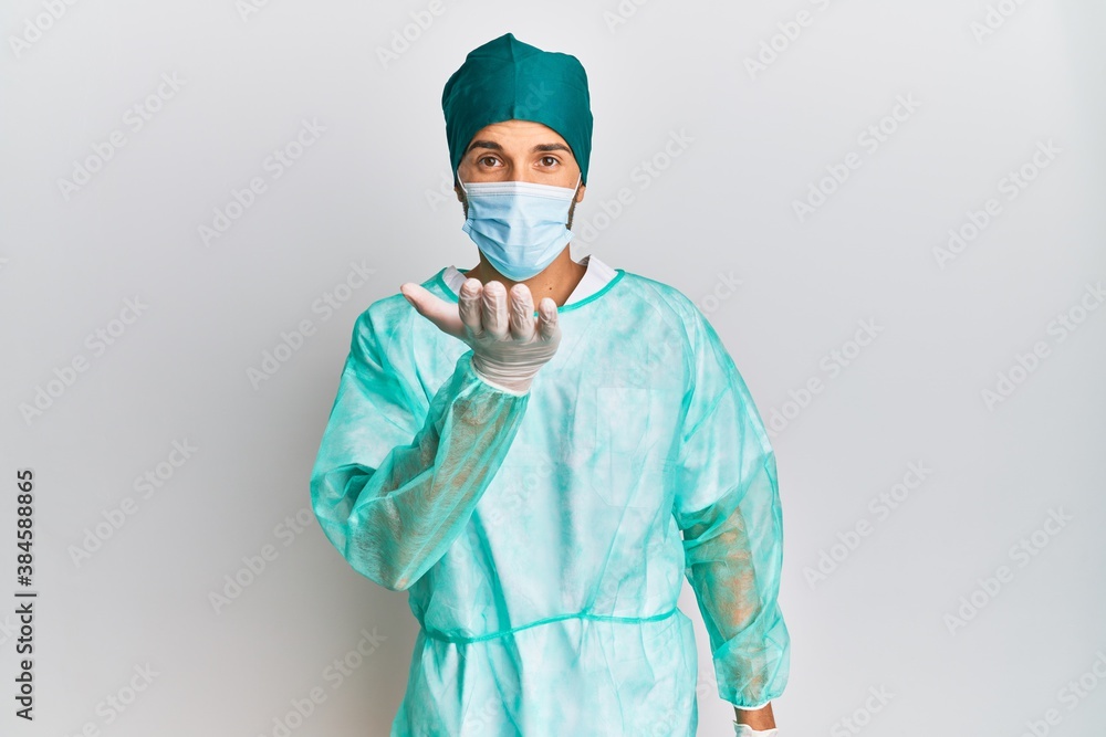 Young handsome man wearing surgeon uniform and medical mask looking at the camera blowing a kiss with hand on air being lovely and sexy. love expression. Young handsome man wearing surgeon uniform and medical mask looking at the camera blowing a kiss with hand on air being lovely and sexy. love expression.