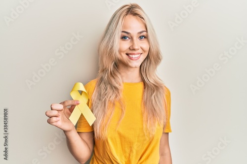 Beautiful caucasian blonde girl holding suicide prevention yellow ribbon looking positive and happy standing and smiling with a confident smile showing teeth