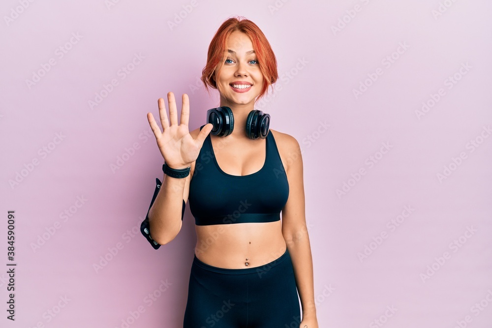 Young beautiful redhead woman wearing gym clothes and using headphones showing and pointing up with fingers number five while smiling confident and happy.