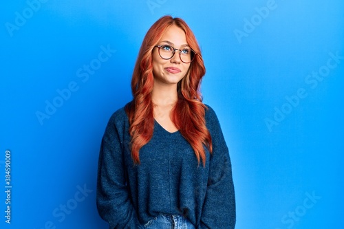 Young beautiful redhead woman wearing casual sweater and glasses over blue background smiling looking to the side and staring away thinking.