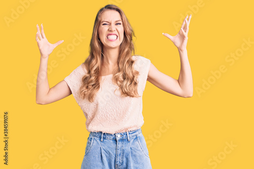 Young beautiful caucasian woman with blond hair wearing casual clothes shouting frustrated with rage, hands trying to strangle, yelling mad