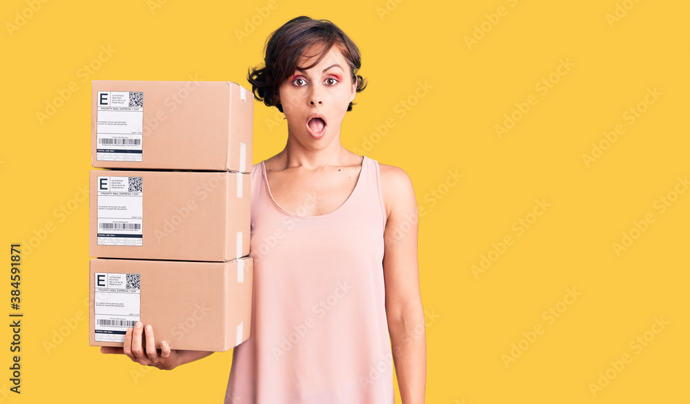 Beautiful young woman with short hair holding delivery packages scared and amazed with open mouth for surprise, disbelief face Beautiful young woman with short hair holding delivery packages scared and amazed with open mouth for surprise, disbelief face