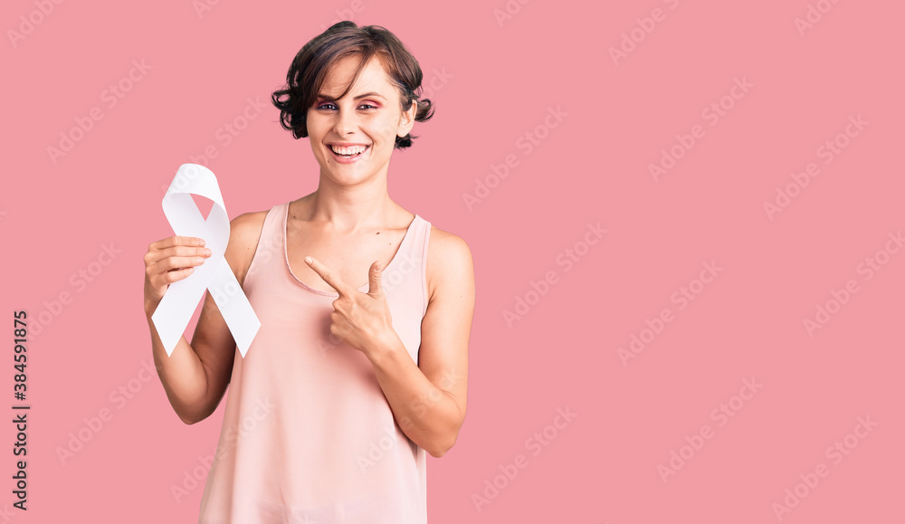 Beautiful young woman with short hair holding cancer awareness white ribbon smiling happy pointing with hand and finger Beautiful young woman with short hair holding cancer awareness white ribbon smiling happy pointing with hand and finger