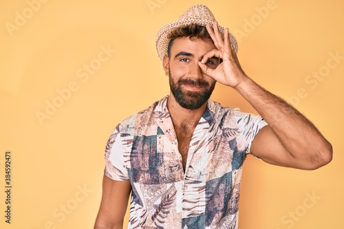 Young hispanic man wearing summer hat doing ok gesture with hand smiling, eye looking through fingers with happy face. © Krakenimages.com