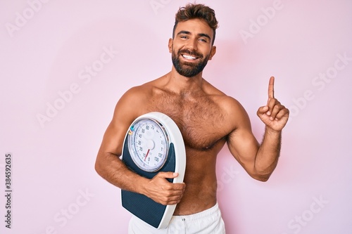 Young hispanic man standing shirtless holding weighing machine surprised with an idea or question pointing finger with happy face  number one