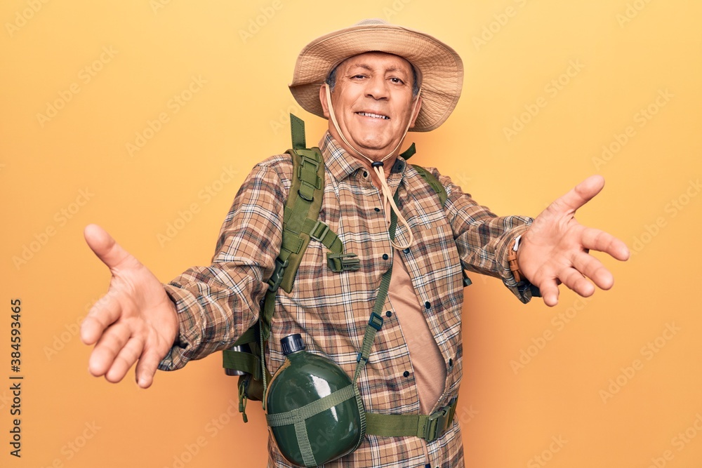 Fototapeta premium Senior man with grey hair wearing hiker backpack and water canteen looking at the camera smiling with open arms for hug. cheerful expression embracing happiness.