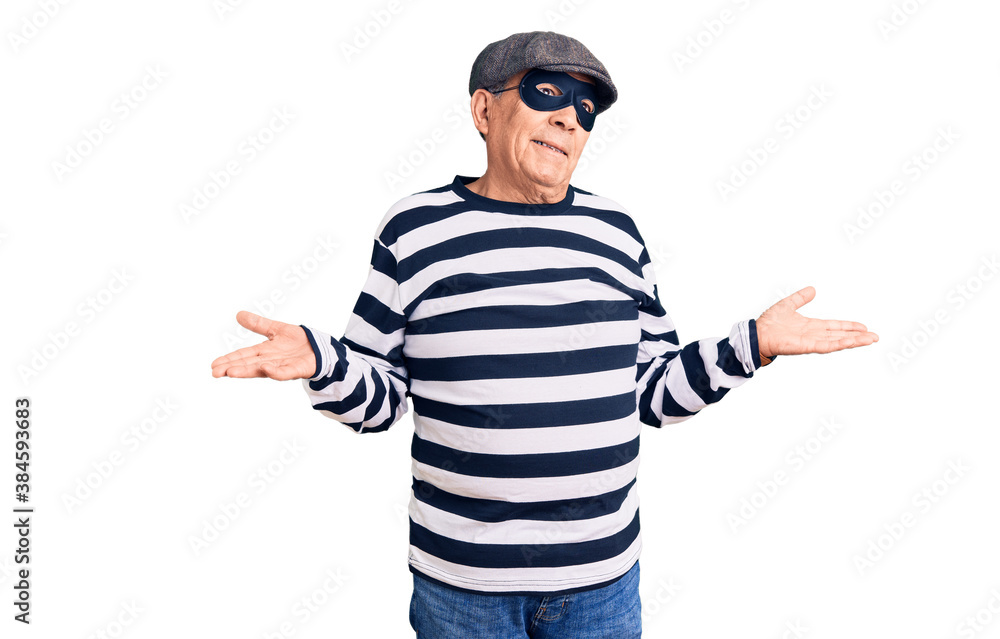 Senior handsome man wearing burglar mask and t-shirt clueless and confused expression with arms and hands raised. doubt concept. Senior handsome man wearing burglar mask and t-shirt clueless and confused expression with arms and hands raised. doubt concept.