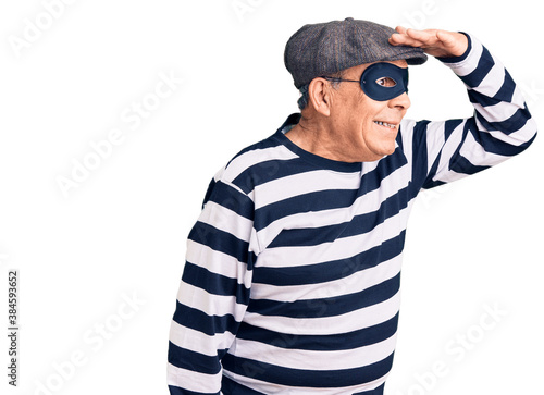 Senior handsome man wearing burglar mask and t-shirt very happy and smiling looking far away with hand over head. searching concept.