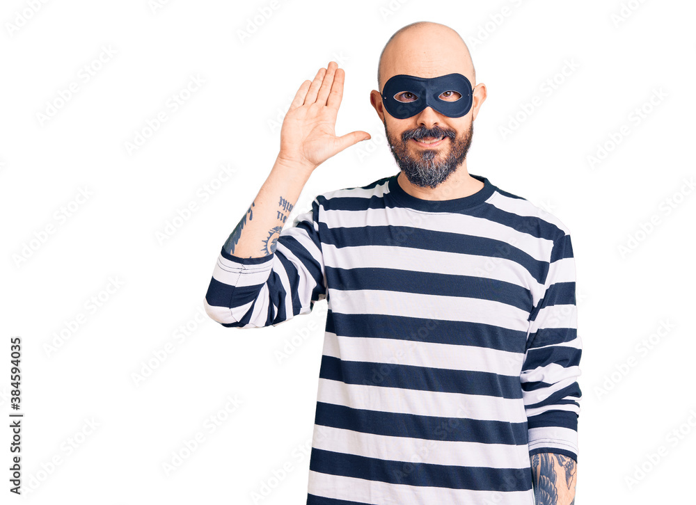 Young handsome man wearing burglar mask waiving saying hello happy and smiling, friendly welcome gesture Young handsome man wearing burglar mask waiving saying hello happy and smiling, friendly welcome gesture