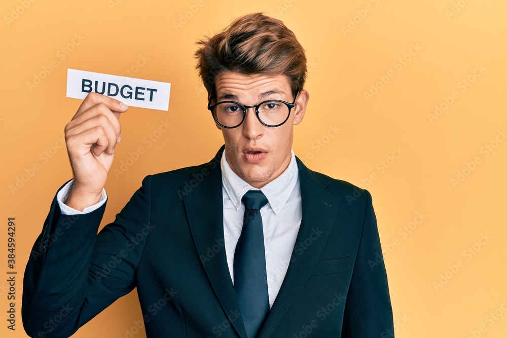 Handsome caucasian business man holding budget word on paper scared and amazed with open mouth for surprise, disbelief face Handsome caucasian business man holding budget word on paper scared and amazed with open mouth for surprise, disbelief face