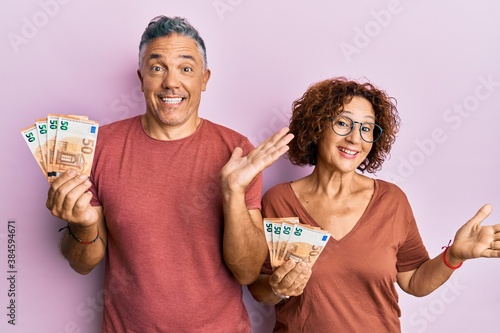Beautiful middle age couple holding bunch of 50 euro banknotes celebrating achievement with happy smile and winner expression with raised hand