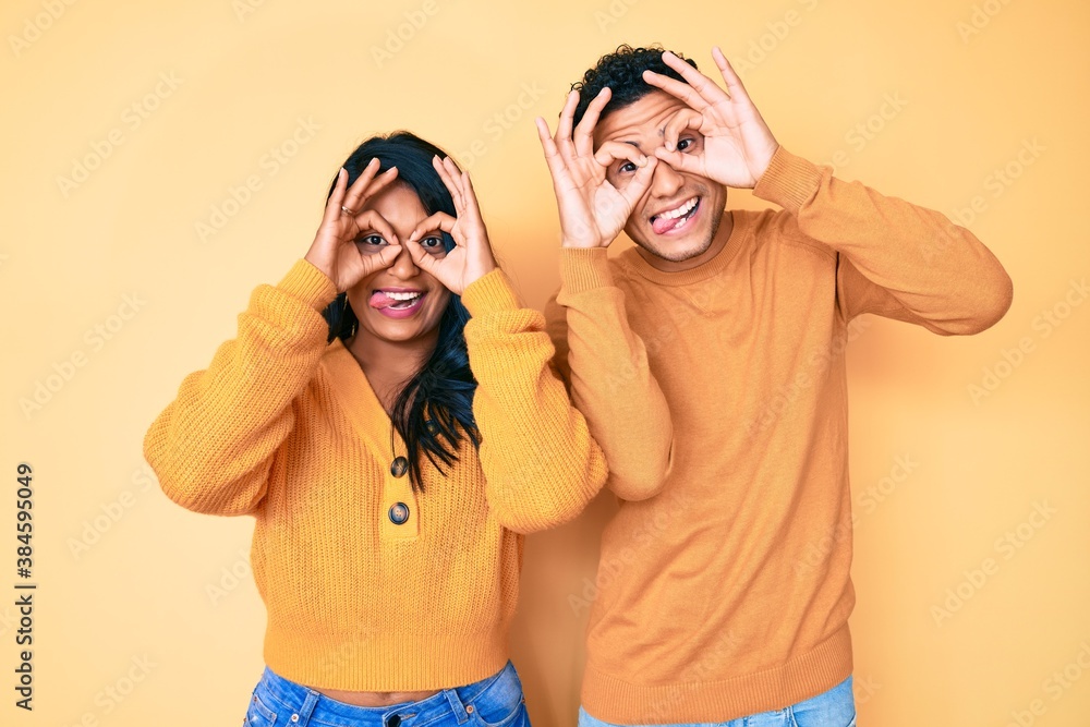 Beautiful latin young couple wearing casual clothes together doing ok gesture like binoculars sticking tongue out, eyes looking through fingers. crazy expression.