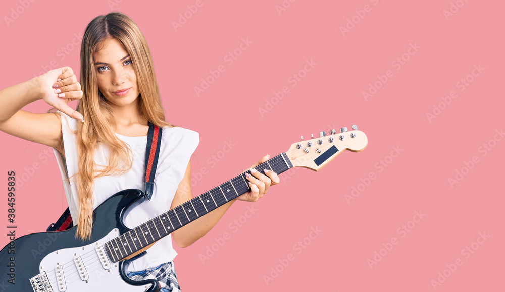 Beautiful caucasian woman with blonde hair playing electric guitar with angry face, negative sign showing dislike with thumbs down, rejection concept Beautiful caucasian woman with blonde hair playing electric guitar with angry face, negative sign showing dislike with thumbs down, rejection concept