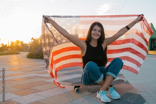 beautiful young woman sitting on a skateboard and holding USA flag in the park at the sunset