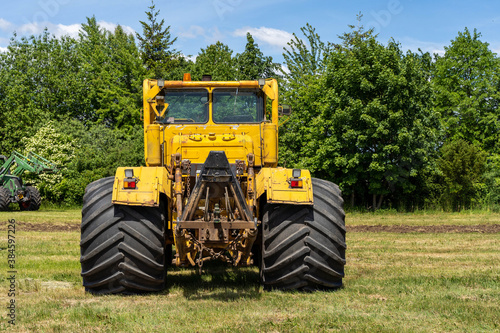 Backside of a yellow tractor with heavy wheels in a field with trees in background