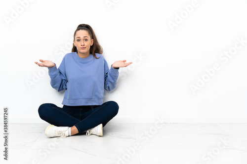 Young woman sitting on the floor having doubts while raising hands © luismolinero
