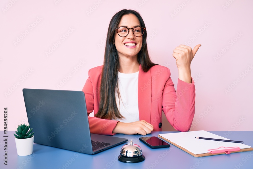 Young caucasian woman sitting at the recepcionist desk working using laptop smiling with happy face looking and pointing to the side with thumb up. Young caucasian woman sitting at the recepcionist desk working using laptop smiling with happy face looking and pointing to the side with thumb up.