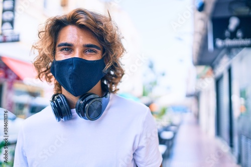 Young handsome hispanic man wearing coronavirus protection mask and using headphones at street of city.