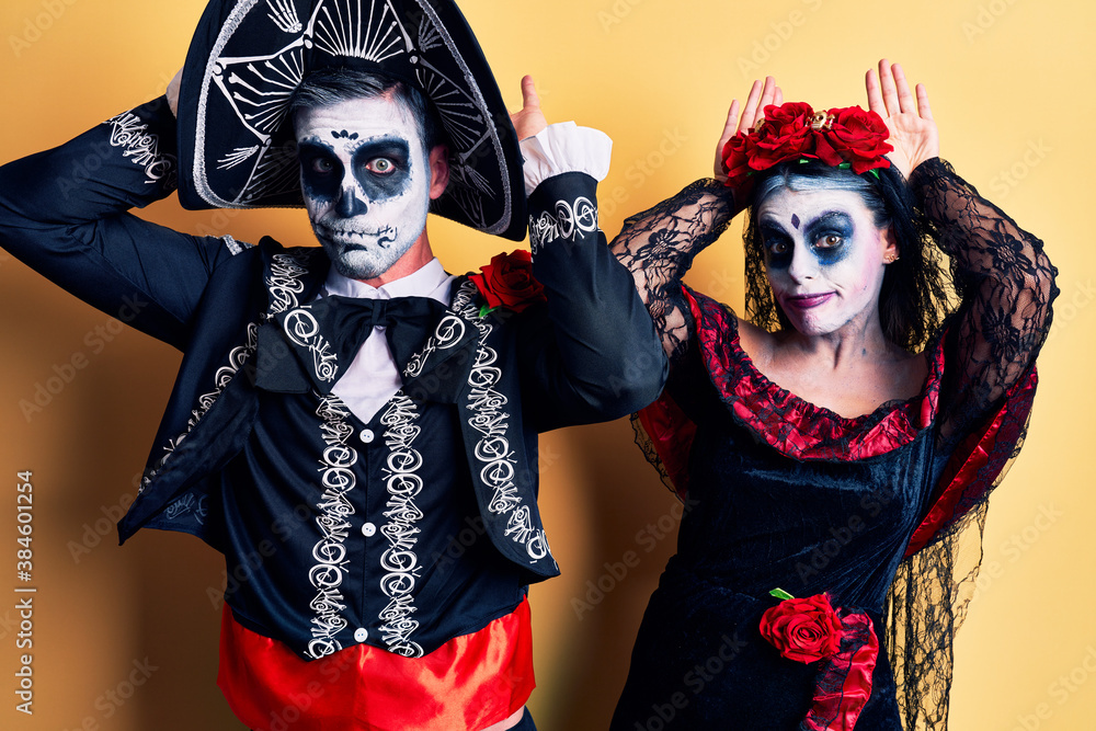 Young couple wearing mexican day of the dead costume over yellow doing bunny ears gesture with hands palms looking cynical and skeptical. easter rabbit concept. Young couple wearing mexican day of the dead costume over yellow doing bunny ears gesture with hands palms looking cynical and skeptical. easter rabbit concept.