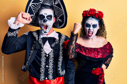 Young couple wearing mexican day of the dead costume over yellow angry and mad raising fist frustrated and furious while shouting with anger. rage and aggressive concept.