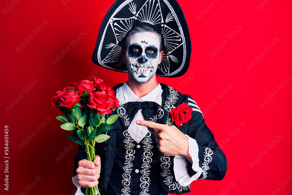 Young man wearing mexican day of the dead costume holding roses smiling happy pointing with hand and finger