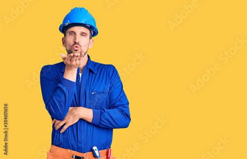Young handsome man wearing worker uniform and hardhat looking at the camera blowing a kiss with hand on air being lovely and sexy. love expression.