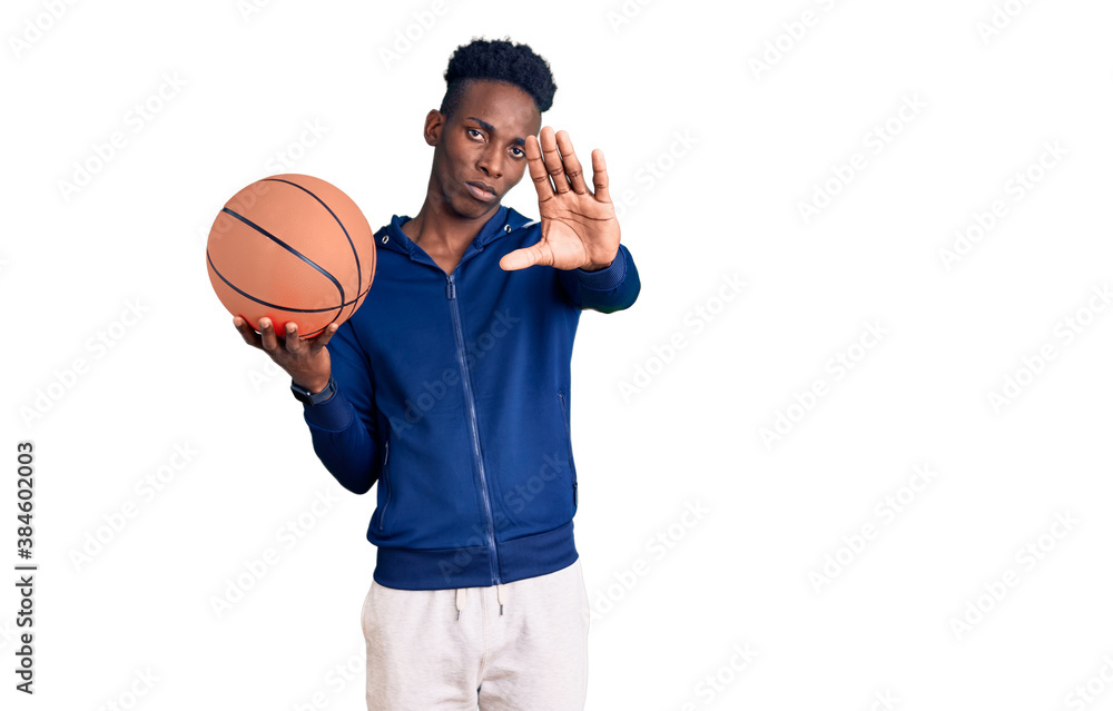 Young african american man holding basketball ball with open hand doing stop sign with serious and confident expression, defense gesture