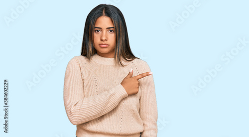 Young latin girl wearing wool winter sweater pointing with hand finger to the side showing advertisement, serious and calm face © Krakenimages.com
