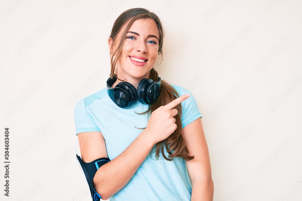 Beautiful young caucasian woman wearing gym clothes and using headphones cheerful with a smile of face pointing with hand and finger up to the side with happy and natural expression on face Beautiful young caucasian woman wearing gym clothes and using headphones cheerful with a smile of face pointing with hand and finger up to the side with happy and natural expression on face