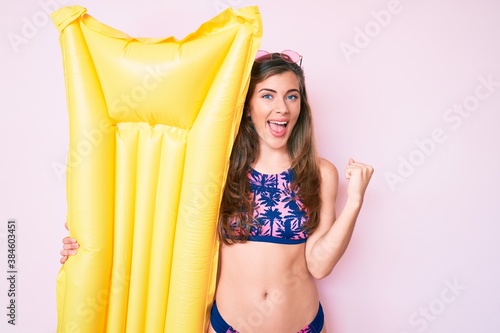 Beautiful young caucasian woman wearing bikini and holding summer matress float screaming proud, celebrating victory and success very excited with raised arms © Krakenimages.com