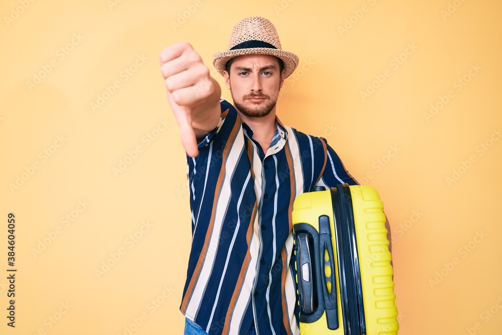 Young handsome man wearing summer hat holding cabin bag with angry face, negative sign showing dislike with thumbs down, rejection concept Young handsome man wearing summer hat holding cabin bag with angry face, negative sign showing dislike with thumbs down, rejection concept