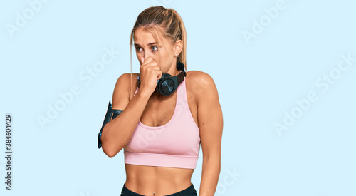 Young blonde woman wearing gym clothes and using headphones smelling something stinky and disgusting, intolerable smell, holding breath with fingers on nose. bad smell