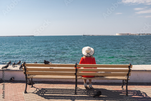 a girl in a red dress and hat sits on a bench in front of the sea and looks at it © John