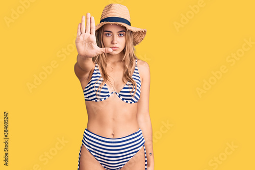 Young beautiful blonde woman wearing bikini and hat doing stop sing with palm of the hand. warning expression with negative and serious gesture on the face.