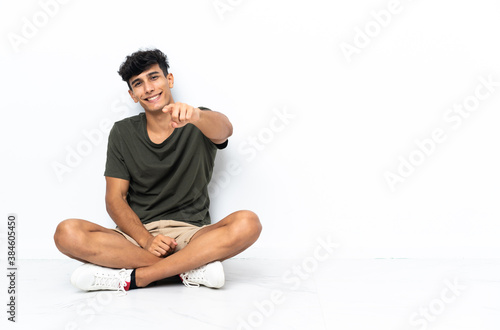 Young Argentinian man sitting on the floor points finger at you with a confident expression