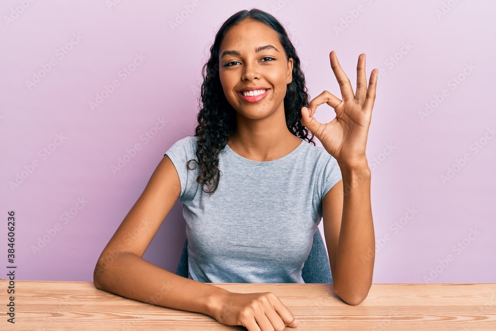 Young african american girl wearing casual clothes sitting on the table smiling positive doing ok sign with hand and fingers. successful expression. Young african american girl wearing casual clothes sitting on the table smiling positive doing ok sign with hand and fingers. successful expression.