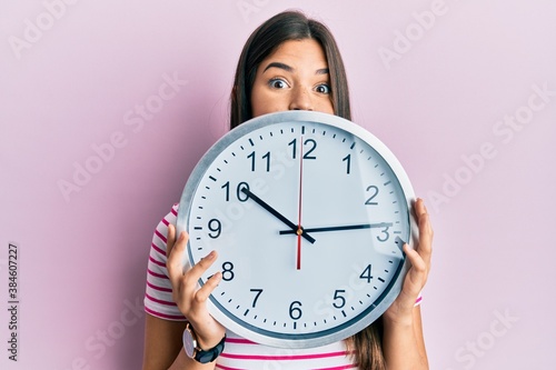 Young brunette woman holding big clock covering face puffing cheeks with funny face. mouth inflated with air, catching air.