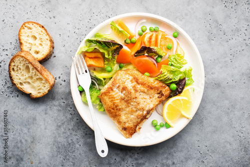 Fried fillet of sea white cod fish with juicy lettuce, capsicum, lemon, green peas on a large white dish on a gray background. Healthy balanced food. Top view,