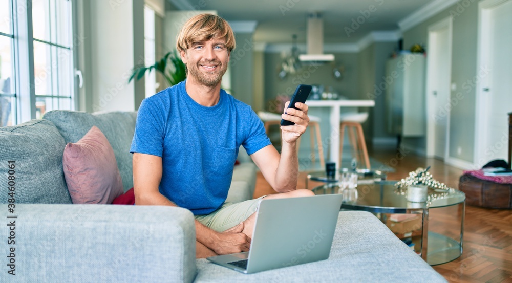 Middle age handsome caucasian man relaxing at home working with computer laptop on the sofa using smartphone Middle age handsome caucasian man relaxing at home working with computer laptop on the sofa using smartphone